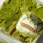 A spoonful of matcha tiramisu lifted from a glass container showing creamy layers of mascarpone, soaked ladyfingers, and vibrant green matcha powder on top.
